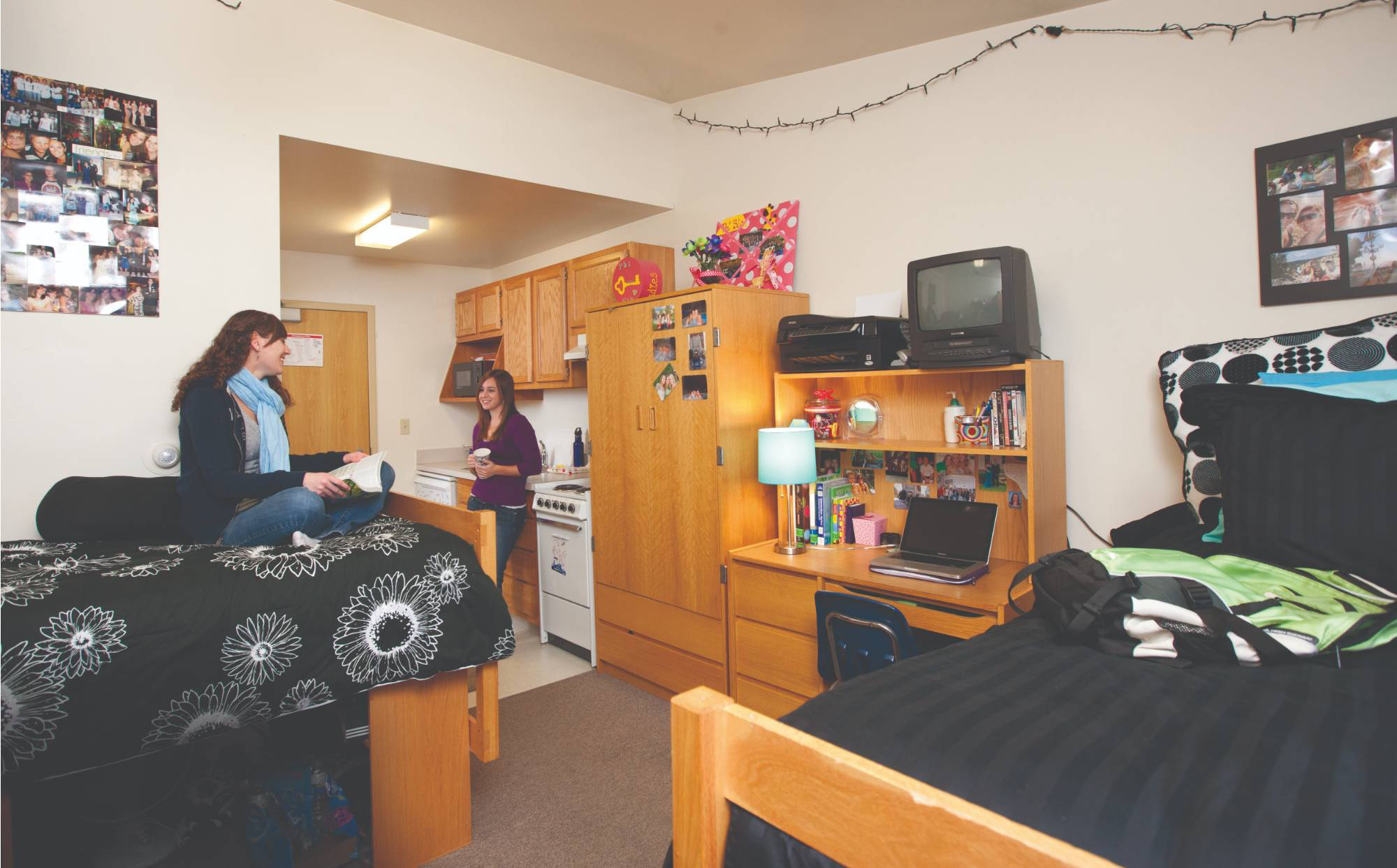 A cozy college 1 bedroom apartment style room with two women chatting. The room has two beds, a small kitchen area, desk, and photos on the walls, conveying a warm, friendly atmosphere.